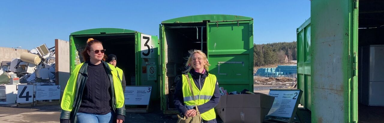 Kaksi opiskelijaa huomioliivit yllään kävelemässä jäteasemalla konttien välissä.
