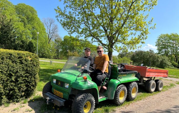 Kaksi puutarha-alan opiskelijaa peräkärryllisen mönkijätraktorin kyydissä, hymyilevät auringonpaisteessa.