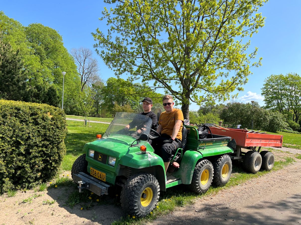 Kaksi puutarha-alan opiskelijaa peräkärryllisen mönkijätraktorin kyydissä, hymyilevät auringonpaisteessa.