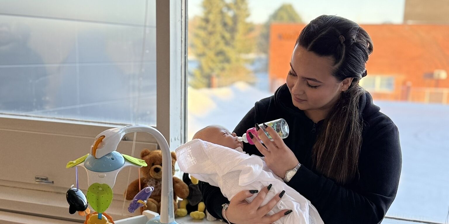 Opiskelija syöttämässä leikkivauvaa tuttipullolla ikkunan edessä.