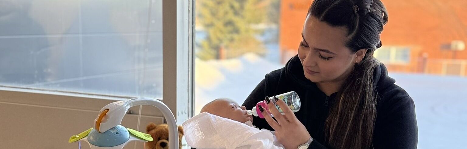 Opiskelija syöttämässä leikkivauvaa tuttipullolla ikkunan edessä.