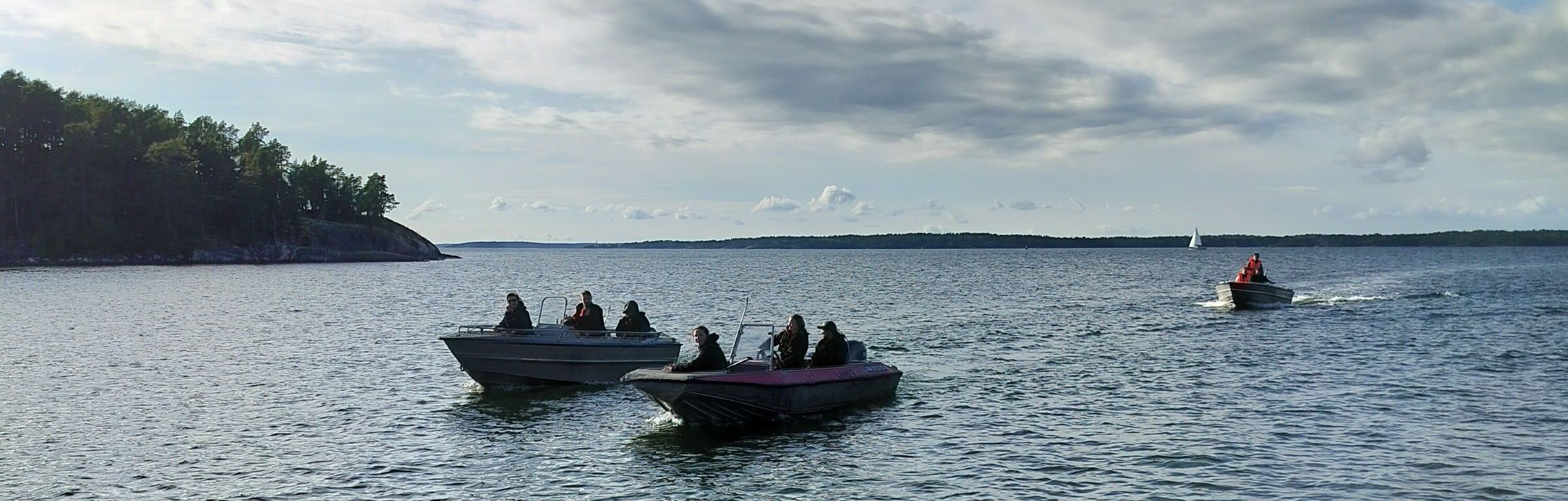 Kolme moottorivenettä matkustajineen merellä, taustalla saari ja avointa merta.